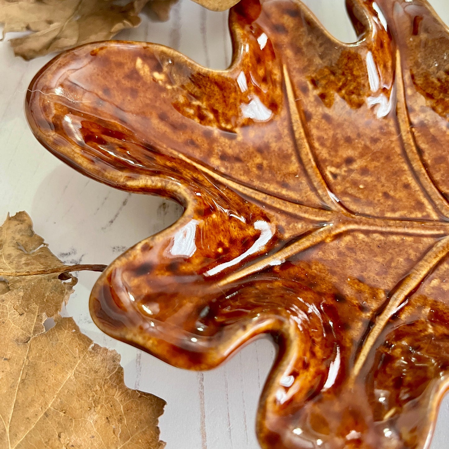 Close up of tip of brown ceramic leaf-shaped dish on a white wooden surface with a dried leaf.
