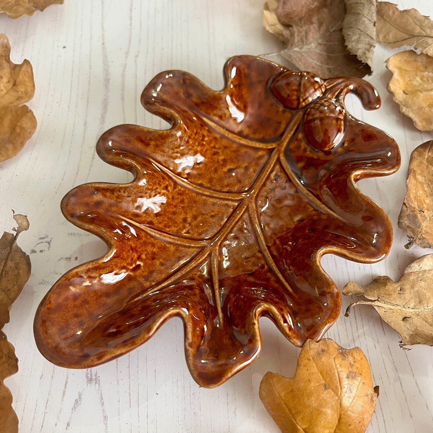 Brown ceramic leaf-shaped dish with acorn on a white surface with scattered leaves.