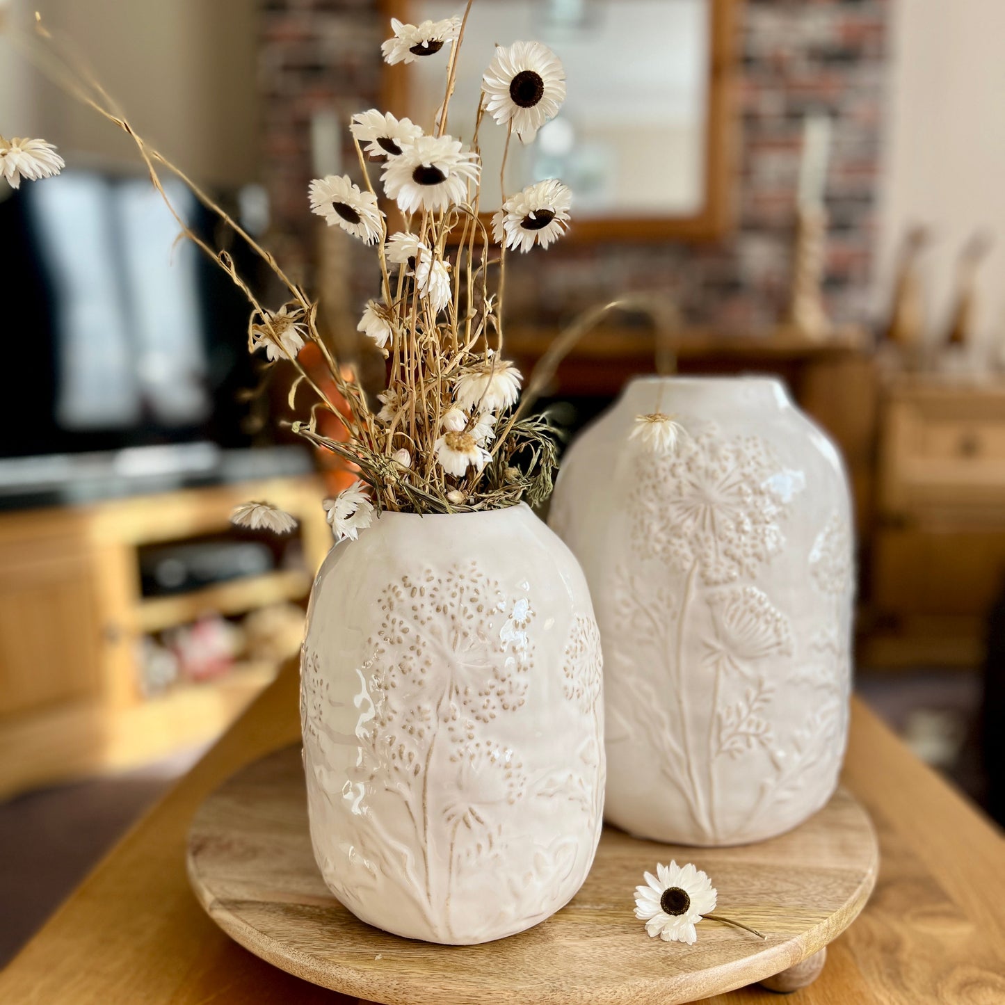 Matching white textured vases with dried flowers on a wooden stand in a living room setting.
