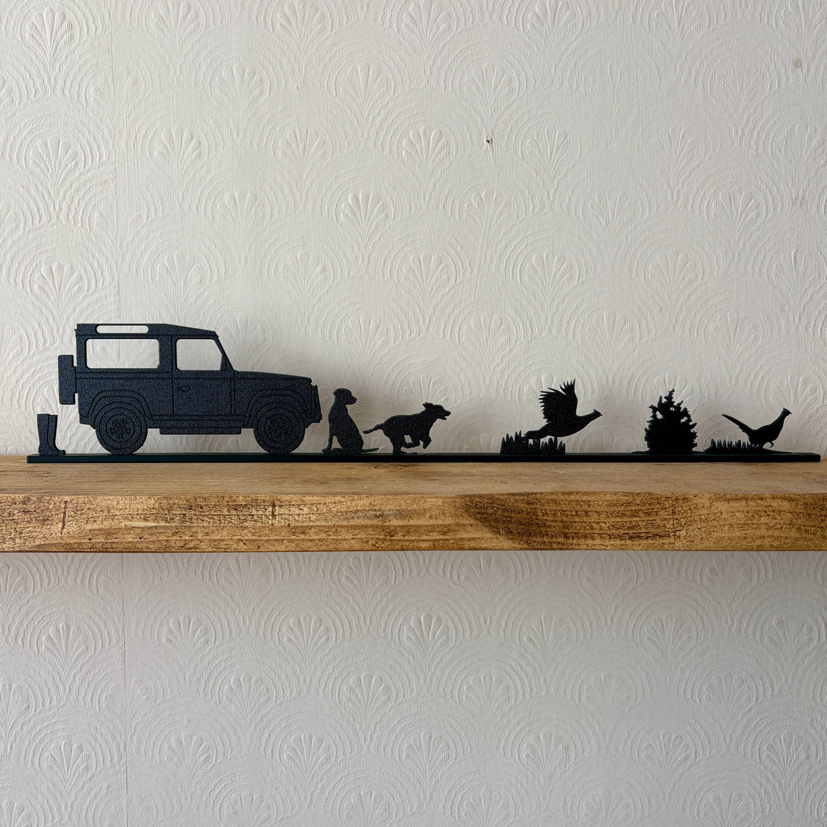 A black shelf topper piece featuring a country scene silhouette set, including a a pair of wellies, Land Rover, Labrador, spaniel & 2 pheasants displayed on a rustic wooden shelf against a white textured wall.