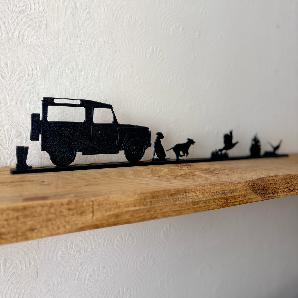 A black shelf topper featuring silhouettes of a Land Rover, a dogs, and pheasants, mounted on a rustic wooden shelf against a white textured wall.