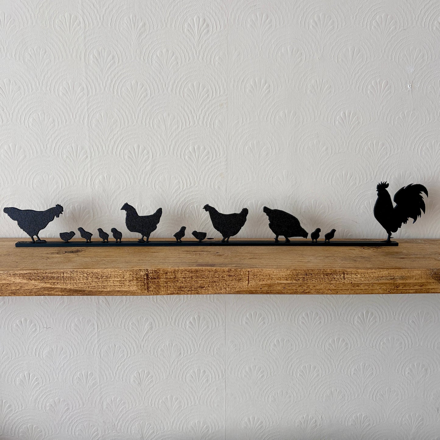 A black recycled plastic topper piece featuring a rooster and several chickens and chicks, displayed on a rustic wooden shelf against a white textured wallpaper background.