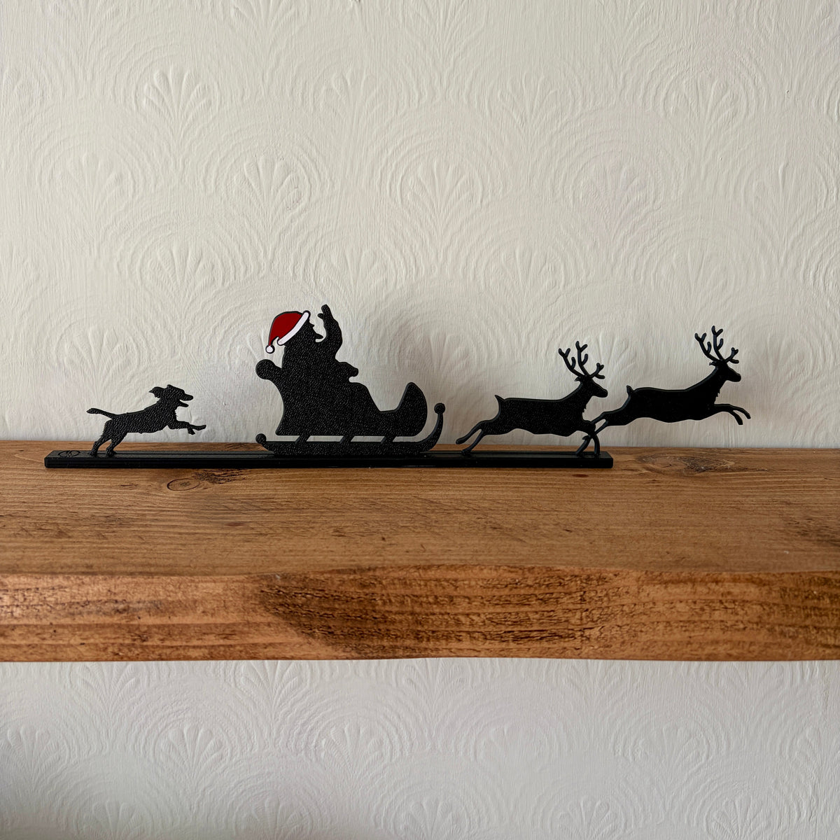 A black recycled plastic Christmas silhouette decoration of Santa Claus in a sleigh pulled by two reindeer and a dog, displayed on a rustic wooden shelf against a textured off-white wall.