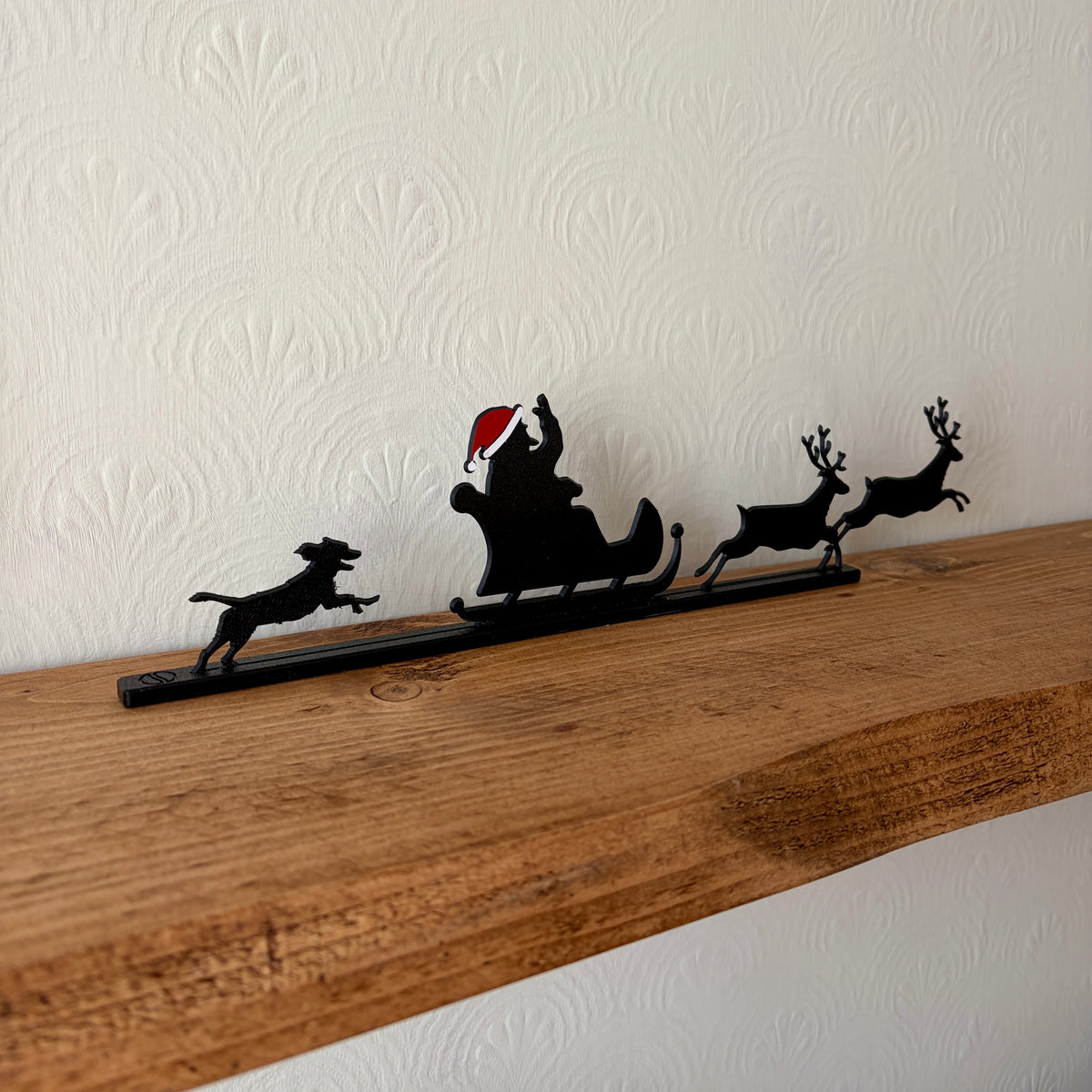 A black recycled plastic Christmas shelf decor piece featuring Santa Claus in a sleigh pulled by three reindeer and a dog, displayed on a wooden mantel against a textured off-white wall.