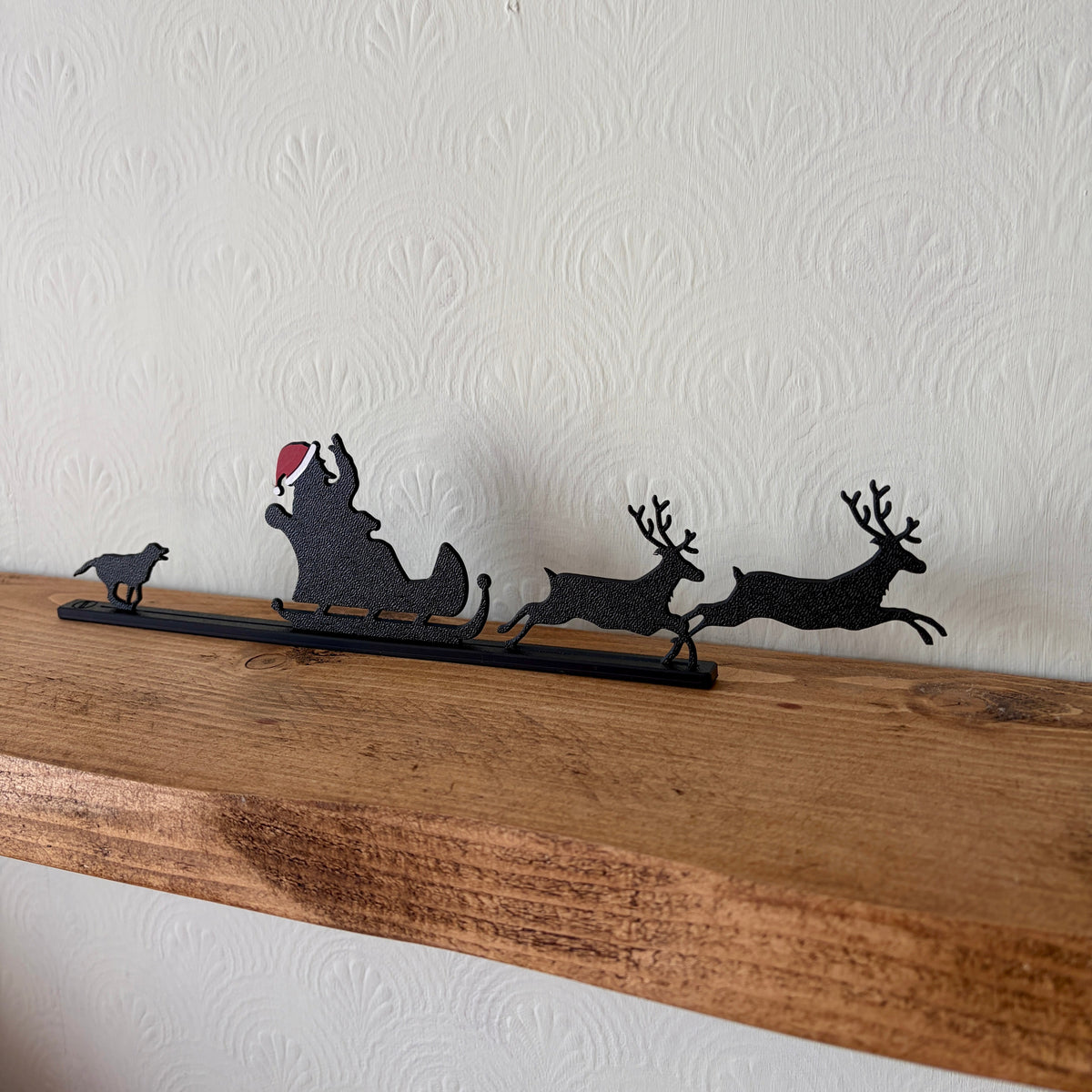 A black recycled plastic silhouette Christmas decor piece featuring Santa Claus in a sleigh pulled by two reindeer, with a small dog on the left, displayed on a rustic wooden shelf against a white textured wall.