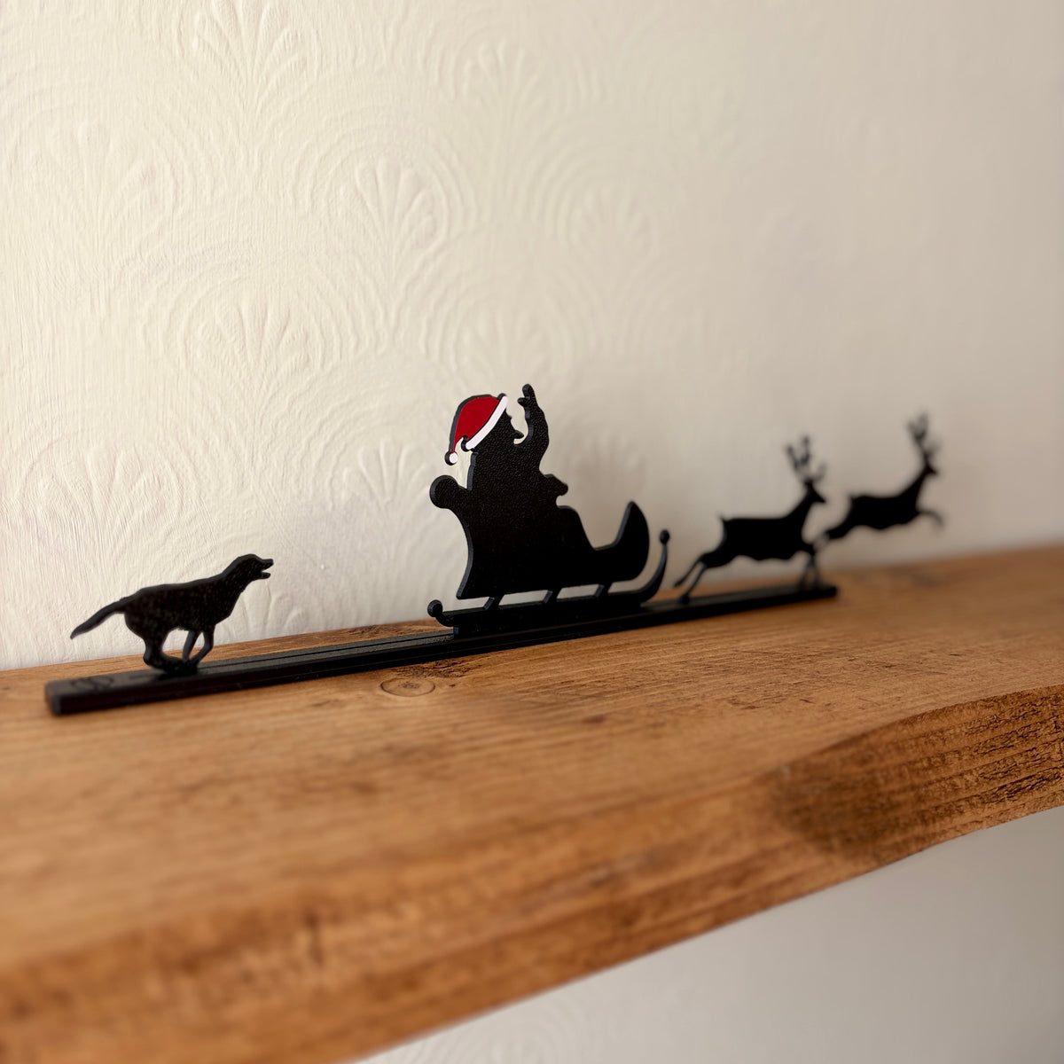 A black recycled plastic Christmas shelf silhouette featuring Santa Claus in a sleigh pulled by two reindeer, with a dog on the left, displayed on a rustic wooden shelf against a textured off-white wall.