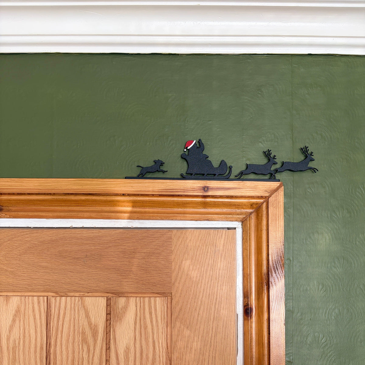 A black recycled plastic silhouette wall decor piece of Santa Claus and his sleigh with reindeer, mounted on a textured olive green wall above a wooden door frame.