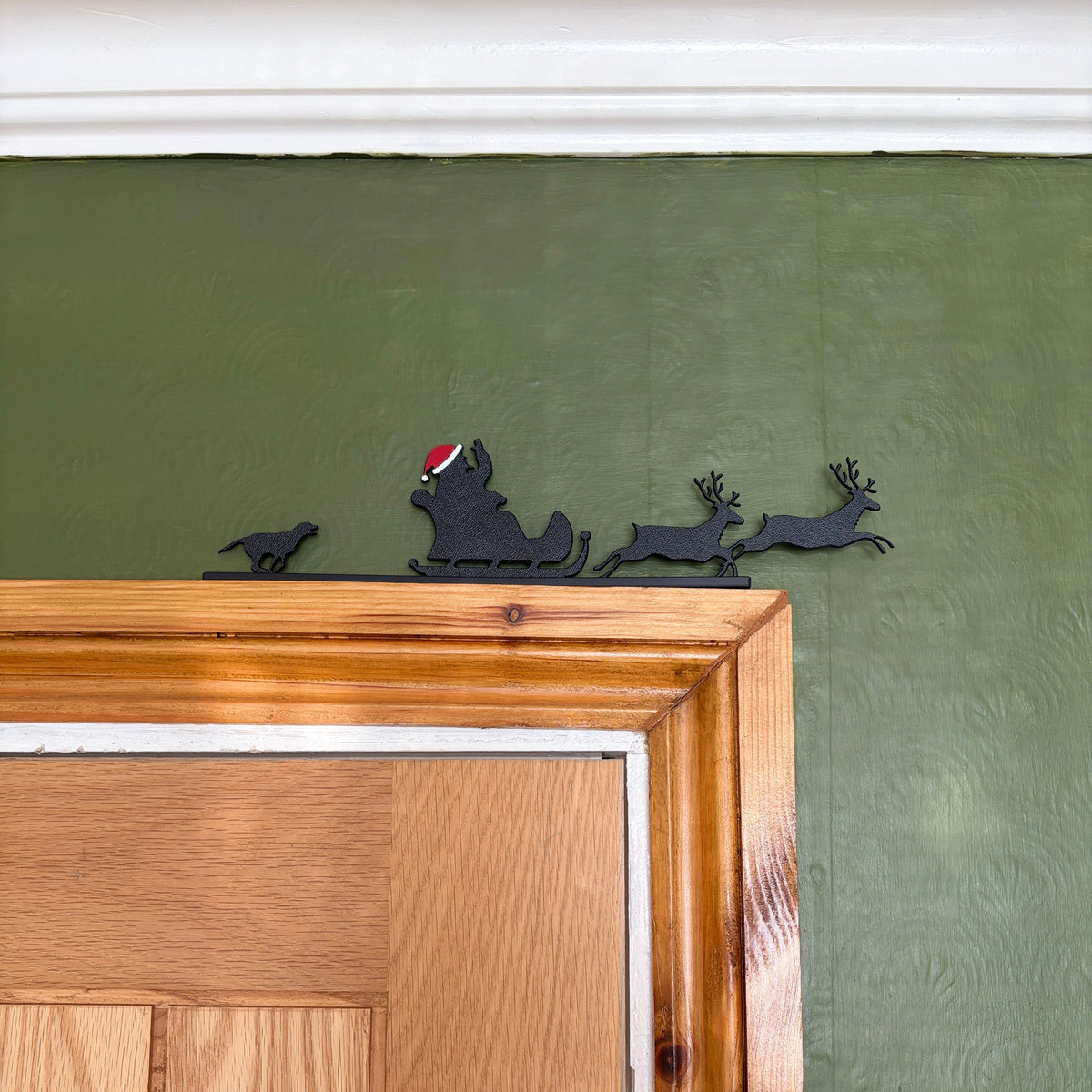 A black recycled plastic decor piece featuring Santa Claus in a sleigh pulled by two reindeer, with a small dog on the left, mounted above a wooden door on a textured olive green wall.
