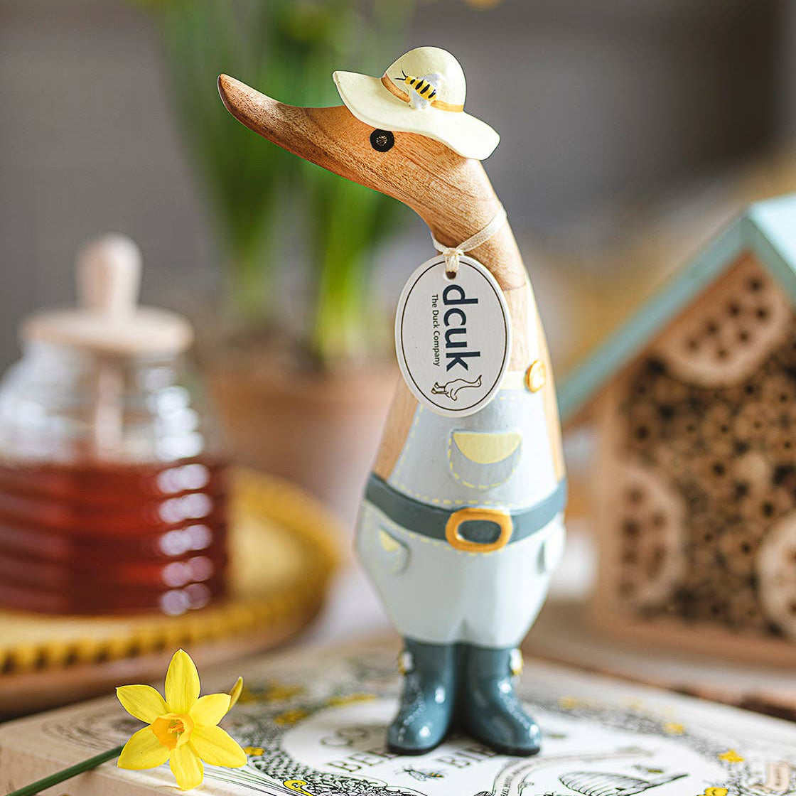 Wooden duck figurine with a hat on a book, surrounded by flowers and a jar of honey.