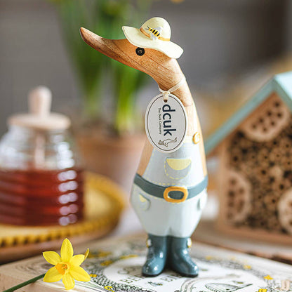 Wooden duck figurine with a hat on a book, surrounded by flowers and a jar of honey.