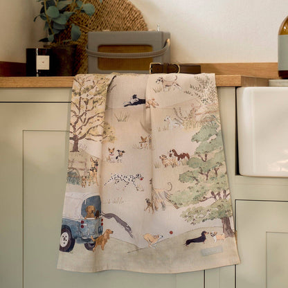 A beige kitchen tea towel with a fun country scene print featuring dogs, trees, & a blue 4x4, hanging over a sage green cabinet in a kitchen setting.