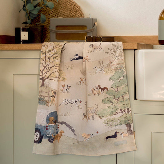 A beige kitchen tea towel with a fun country scene print featuring dogs, trees, & a blue 4x4, hanging over a sage green cabinet in a kitchen setting.
