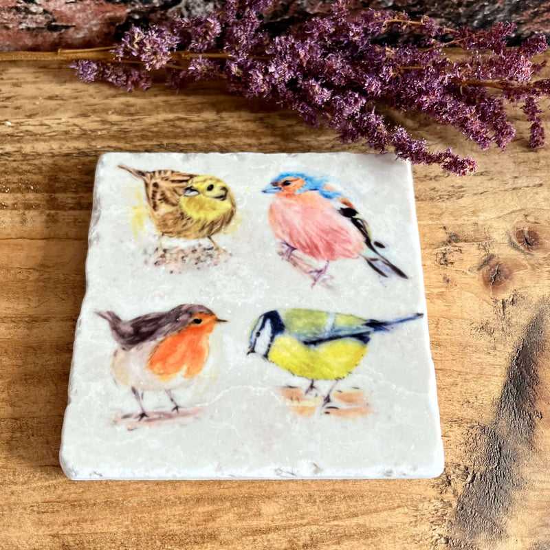 A square natural marble coaster with 4 watercolour painted garden birds displayed on a shelf with purple dried flowers as decoration.