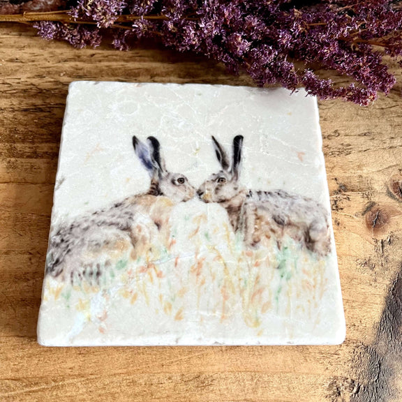 A square natural marble coaster with 2 watercolour painted hares kissing, displayed on a shelf with purple dried flowers as decoration.