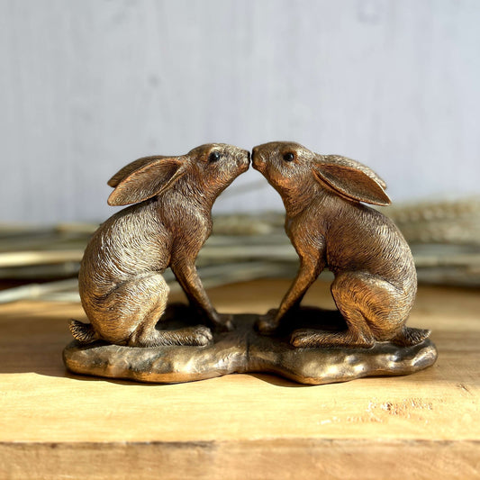 Gold Coloured Hares Ornament Kissing One an Other with Dried Wheat in the Background on a Displayed on a Wooden Shelf.