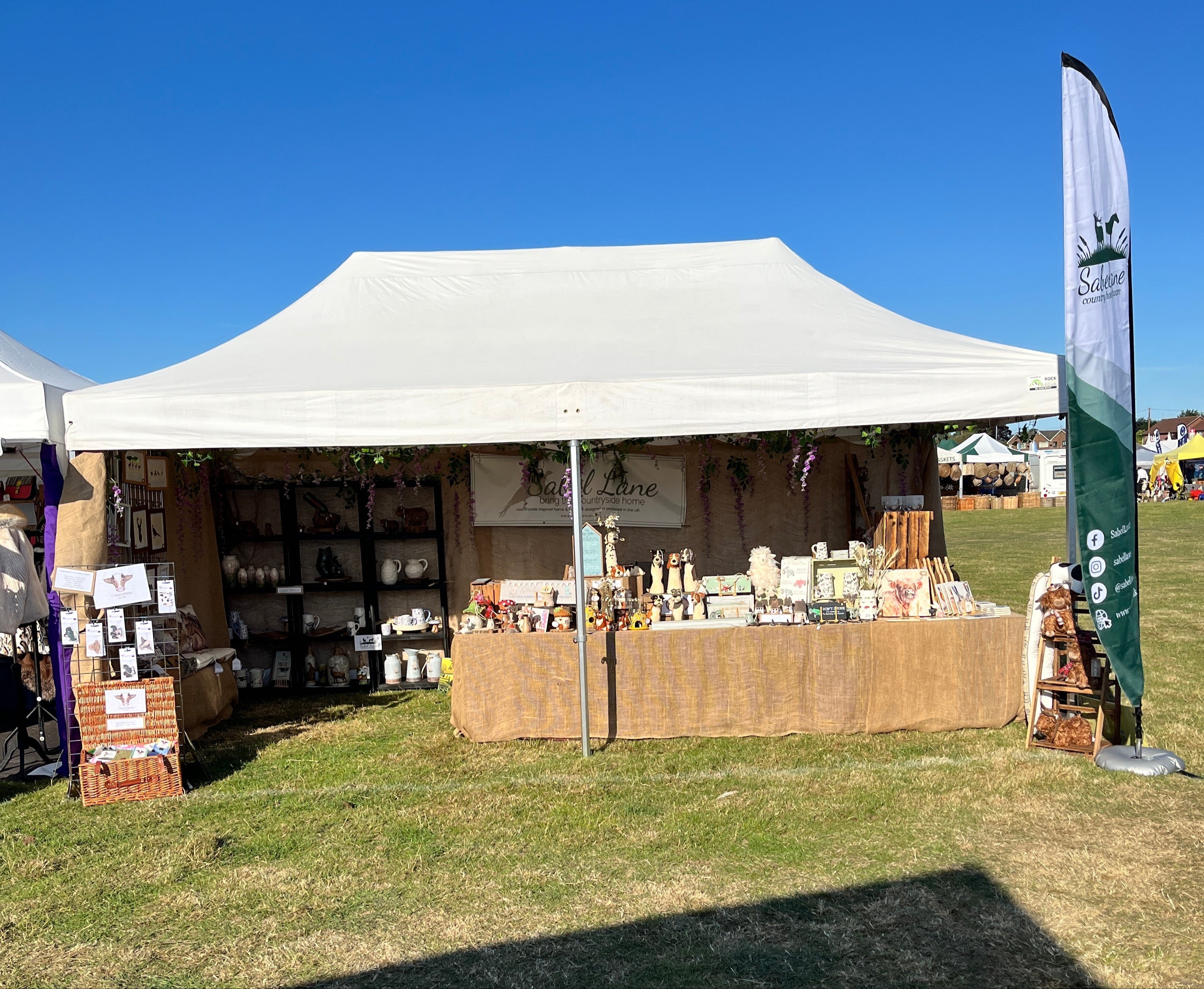 Outdoor market stall with products under a white tent on a grassy area.