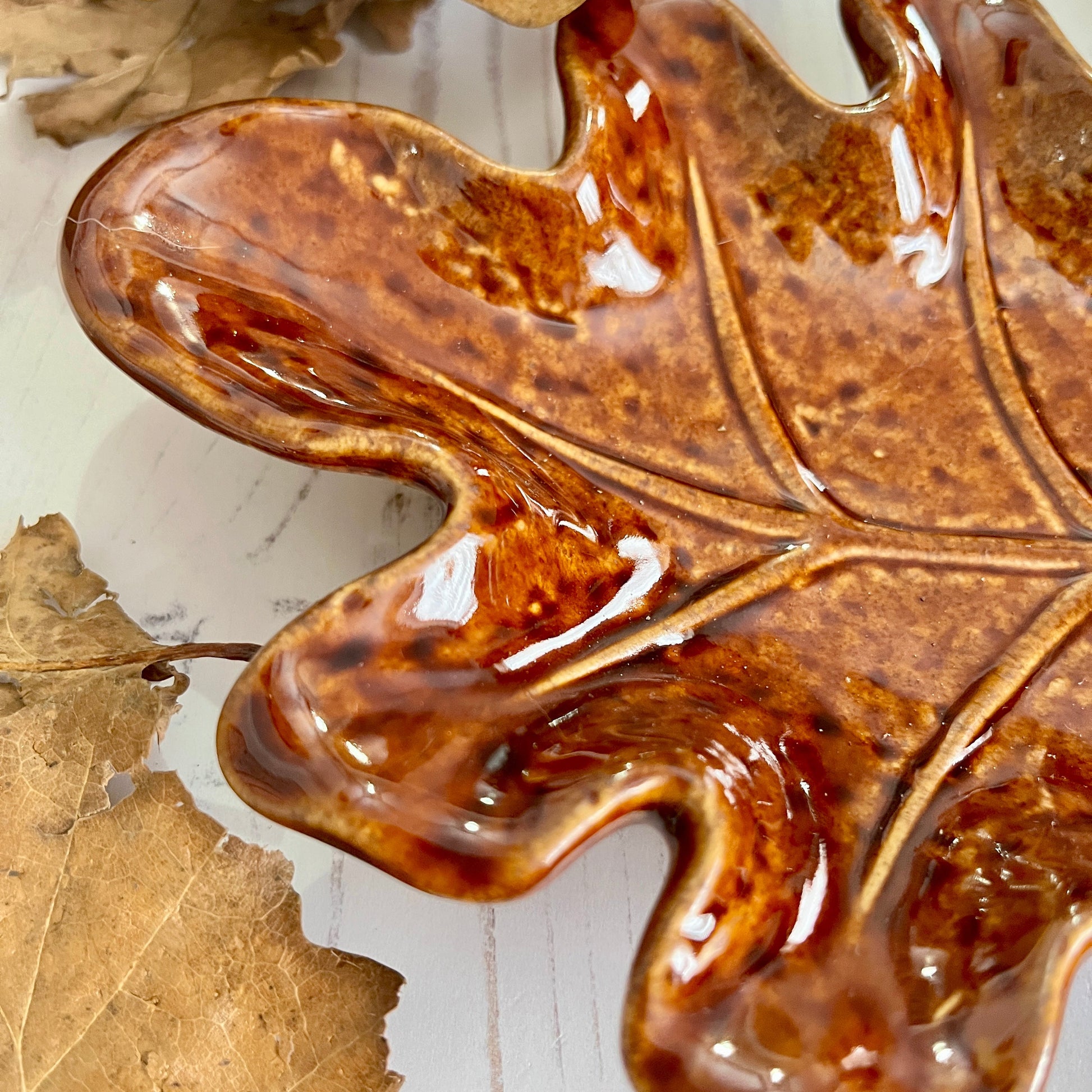 Close up of tip of brown ceramic leaf-shaped dish on a white wooden surface with a dried leaf.