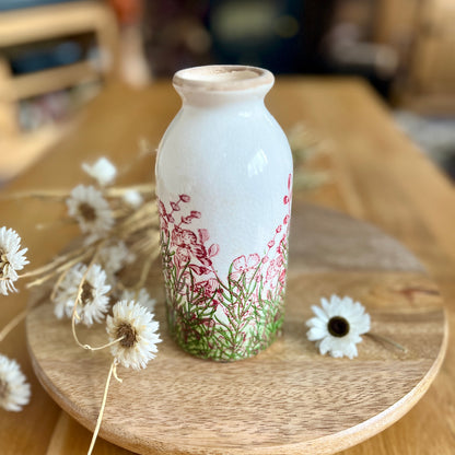 Decorative bottle shaped vase with floral design on a wooden stand with flowers around