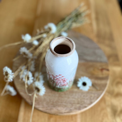 Small white bottle shaped vase with pink floral patterns on a wooden surface with dried plants
