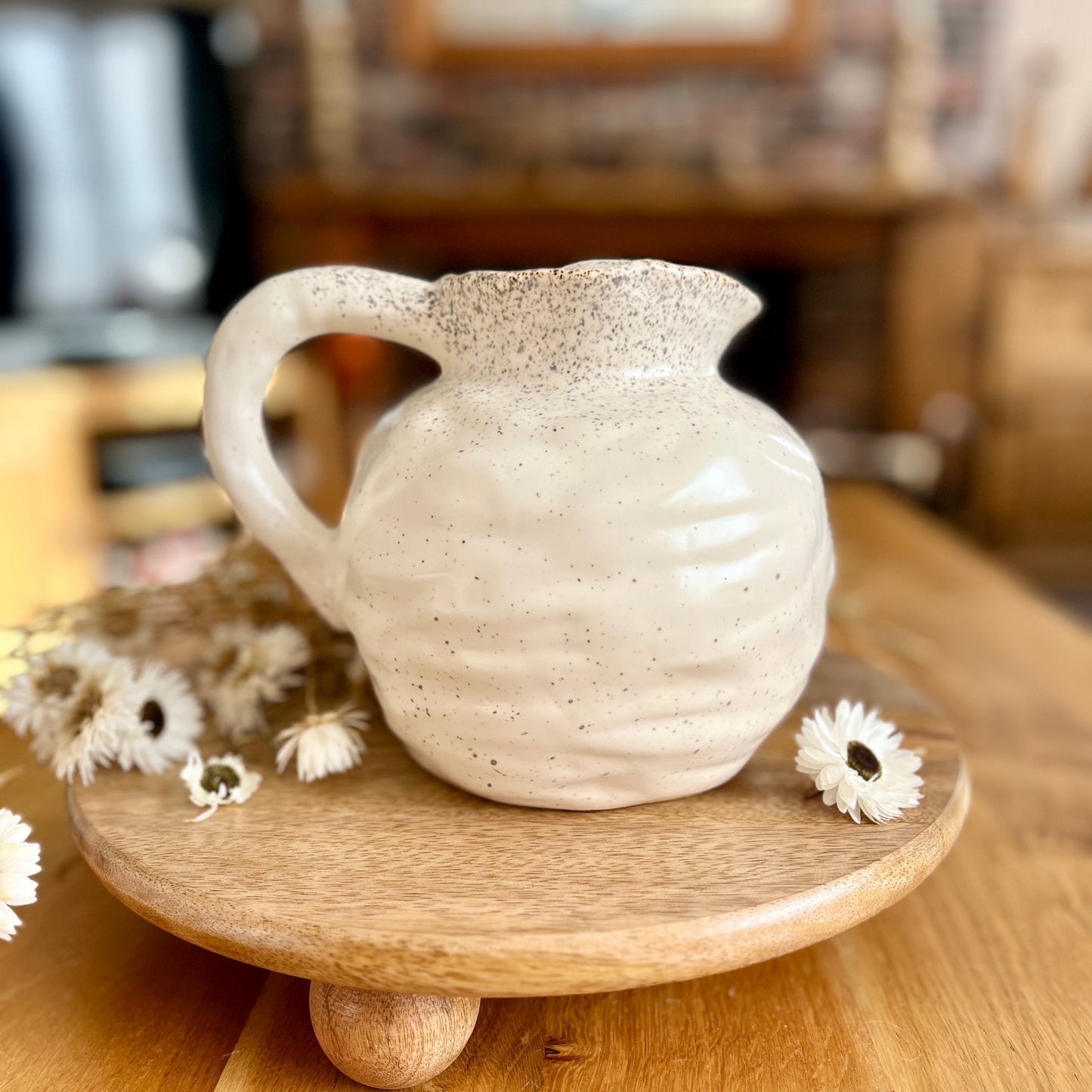 White round ceramic jug on a wooden stand with flowers in a blurred indoor setting