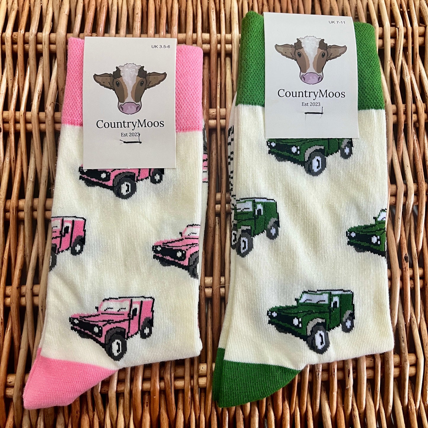 Two pairs of socks with Land Rover patterns (one pink, one green) on a wicker surface, featuring 'Country Moos' branding.