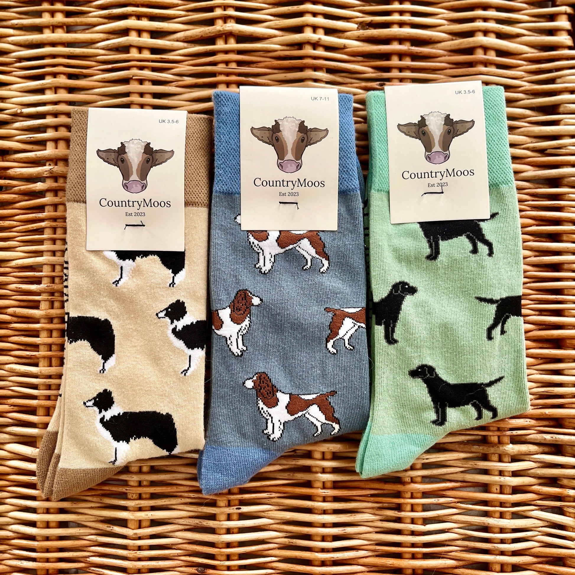 Three pairs of socks with dog patterns on a wicker surface, featuring 'Country Moos' branding.