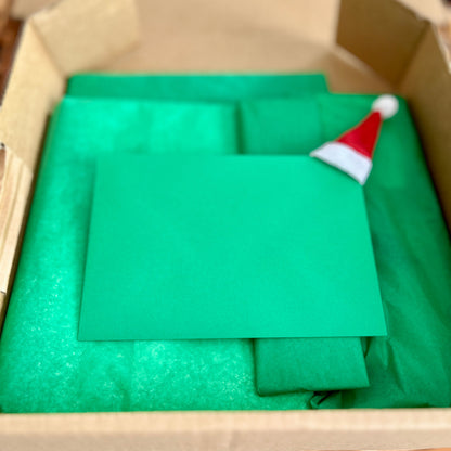 Green paper with a Santa hat on top of it inside an open cardboard box.