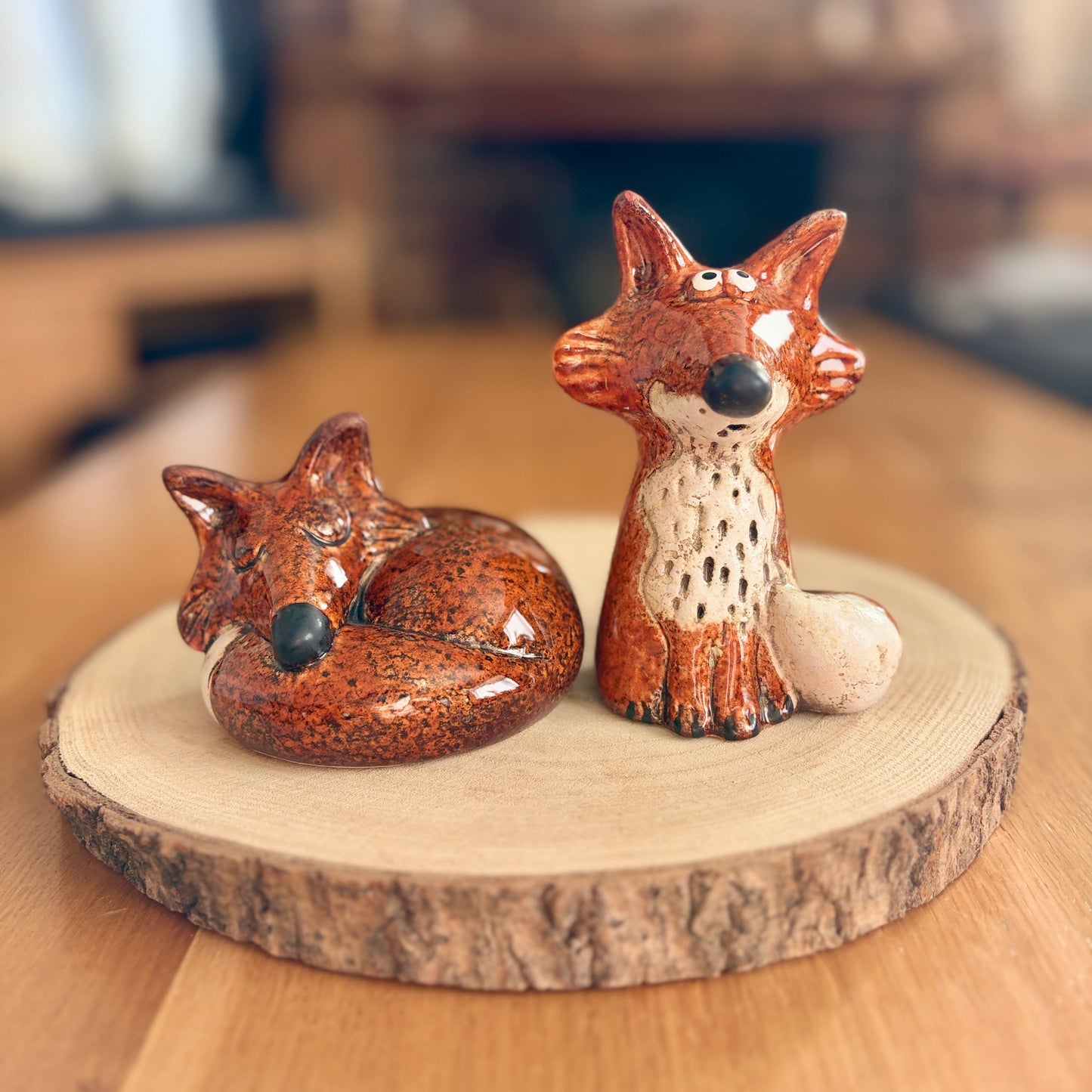 Two ceramic fox figurines on a wooden base with a blurred indoor background