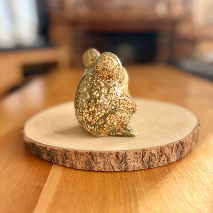 Decorative frog figurine from behind on a wooden base with a blurred indoor background