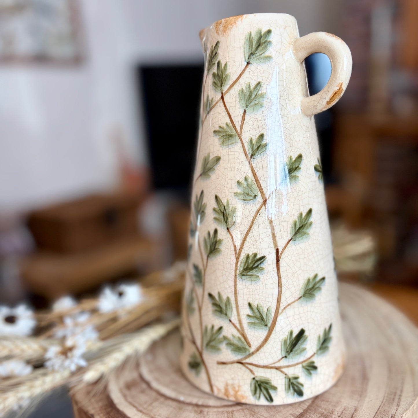 Handle view of a ceramic jug with painted leaves pattern sat on a stool in a home setting.
