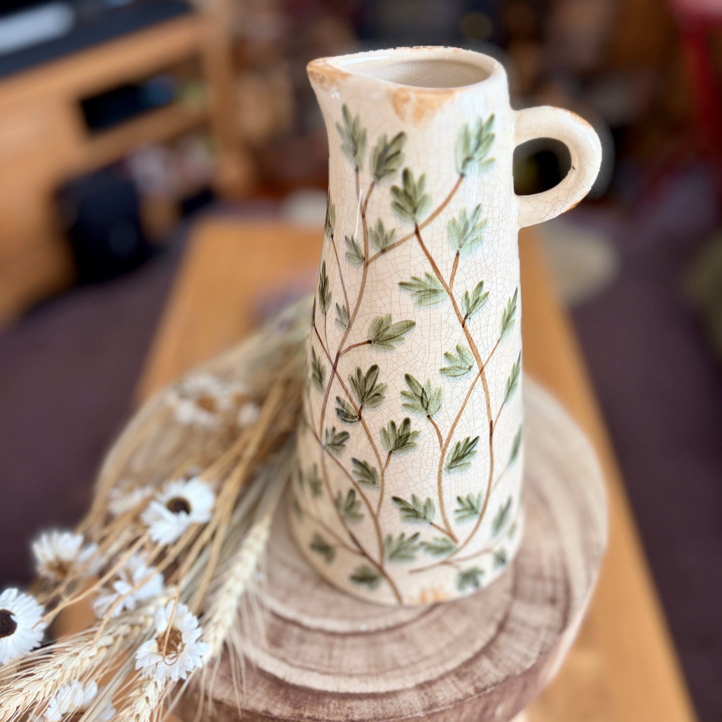 View of the top of a ceramic jug that is decorated with a leafy pattern. It is sat on a wooden stool in a home setting with dried flowers next to it.