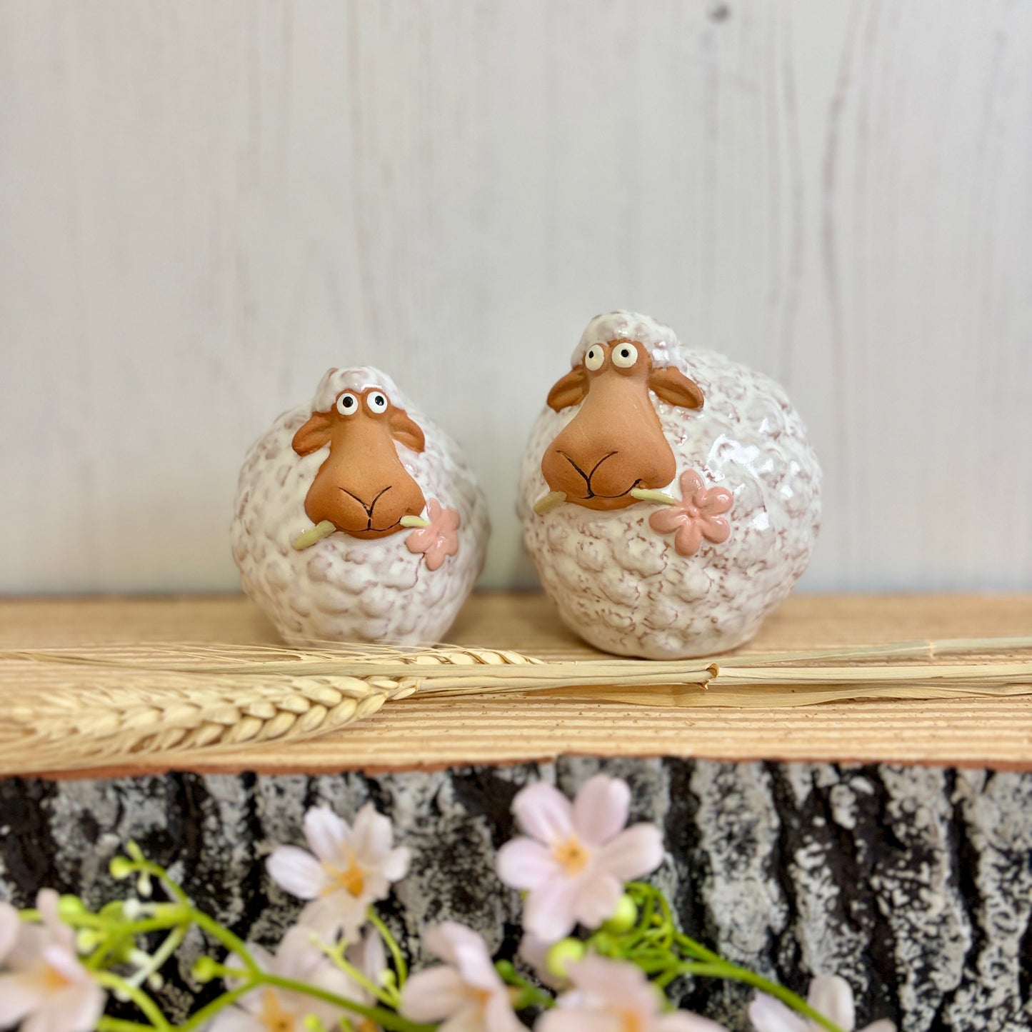 2 ceramic sheep figurines on wooden plinth with pink blossom, wheat and white background