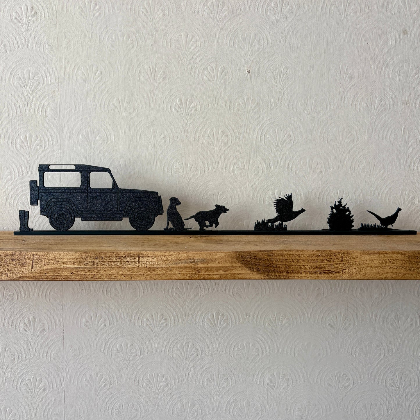 A black shelf topper piece featuring a country scene silhouette set, including a a pair of wellies, Land Rover, Labrador, spaniel & 2 pheasants displayed on a rustic wooden shelf against a white textured wall.