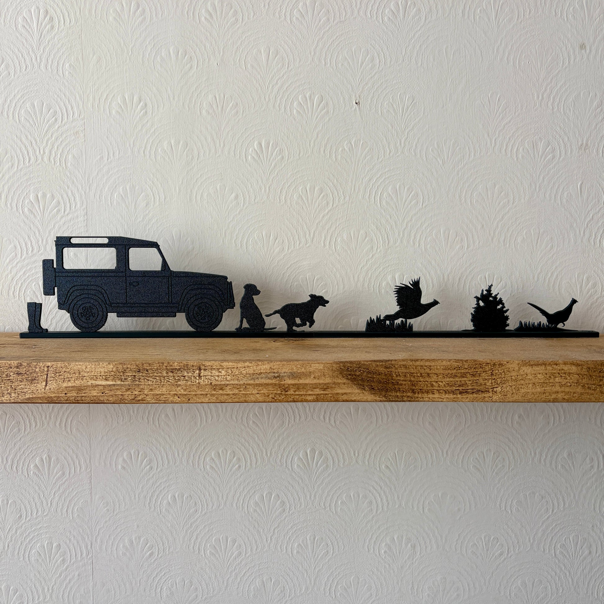 A black shelf topper piece featuring a country scene silhouette set, including a a pair of wellies, Land Rover, Labrador, spaniel & 2 pheasants displayed on a rustic wooden shelf against a white textured wall.