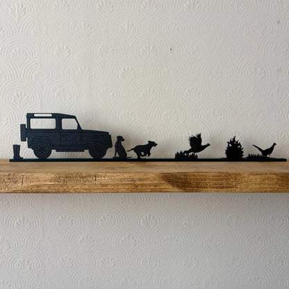 A black shelf topper piece featuring a country scene silhouette set, including a a pair of wellies, Land Rover, Labrador, spaniel & 2 pheasants displayed on a rustic wooden shelf against a white textured wall.