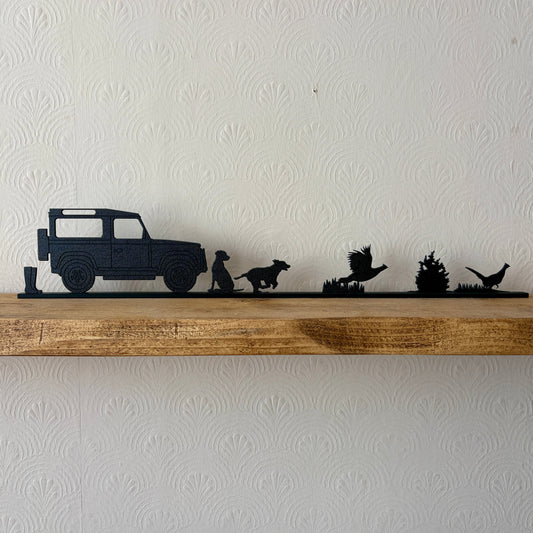 A black shelf topper piece featuring a country scene silhouette set, including a a pair of wellies, Land Rover, Labrador, spaniel & 2 pheasants displayed on a rustic wooden shelf against a white textured wall.