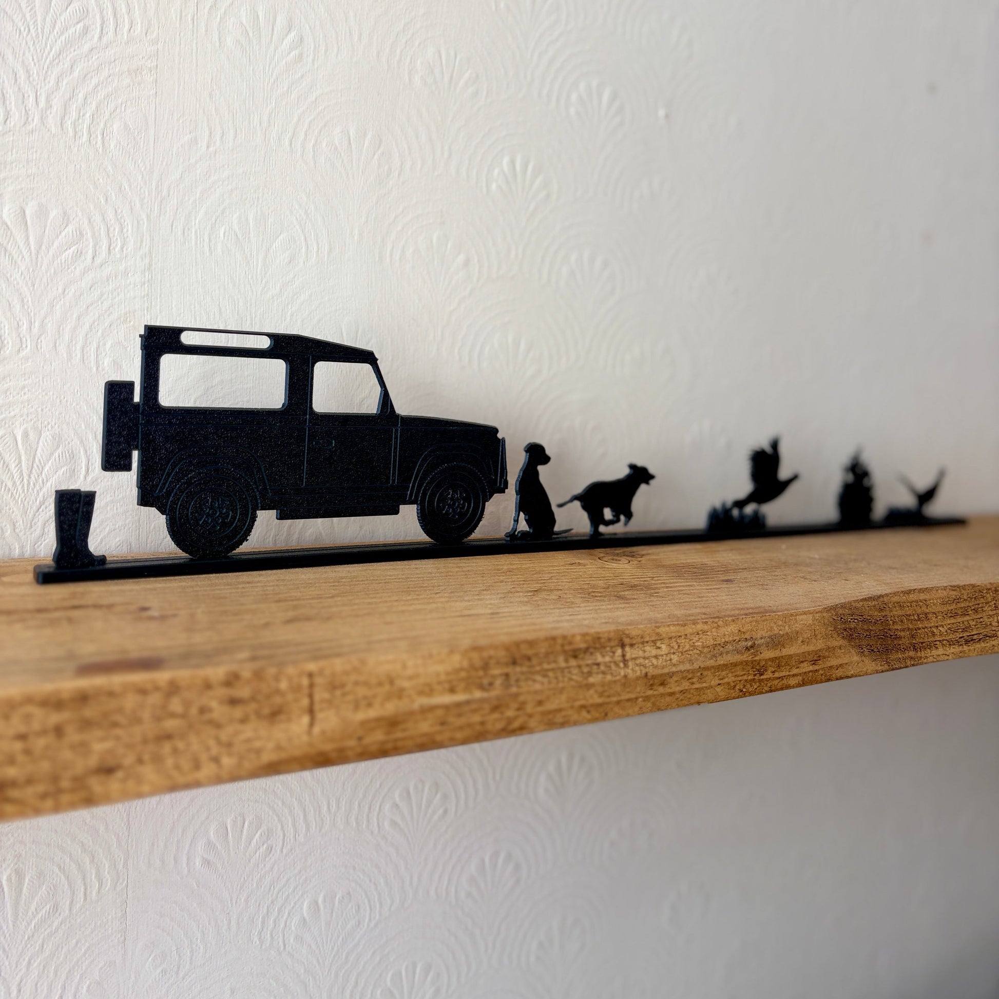 A black shelf topper featuring silhouettes of a Land Rover, a dogs, and pheasants, mounted on a rustic wooden shelf against a white textured wall.