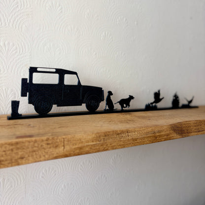 A black shelf topper featuring silhouettes of a Land Rover, a dogs, and pheasants, mounted on a rustic wooden shelf against a white textured wall.