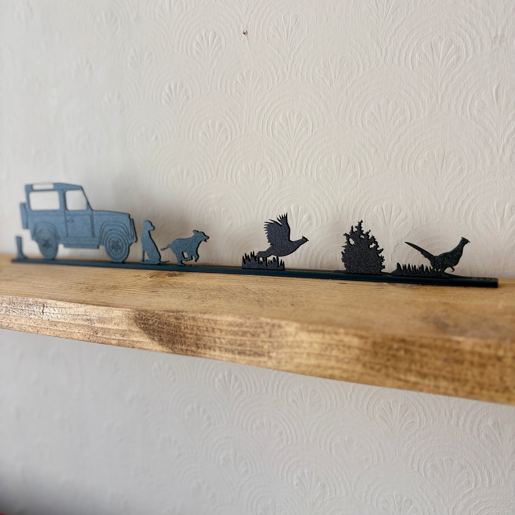A rustic shelf decoration featuring silhouettes of a car, dogs, and pheasants on a black rail, sat on a light wood shelf against a textured off-white wall.