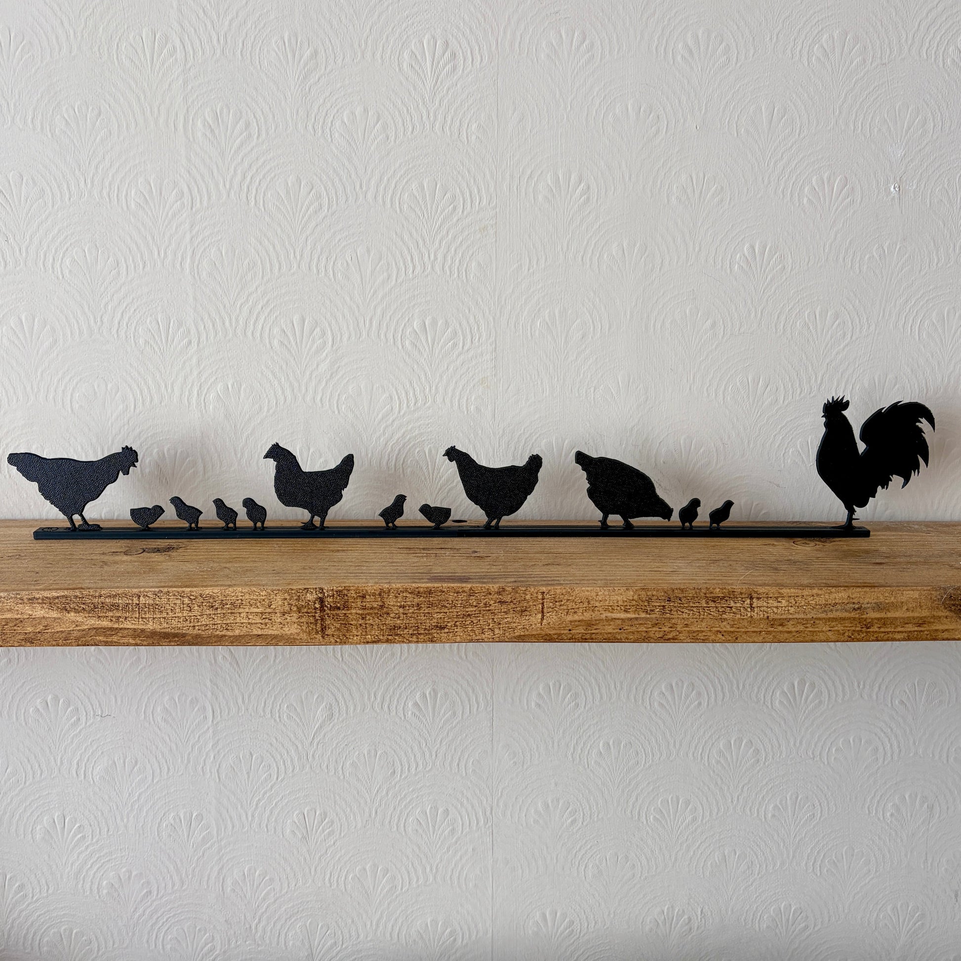 A black recycled plastic topper piece featuring a rooster and several chickens and chicks, displayed on a rustic wooden shelf against a white textured wallpaper background.