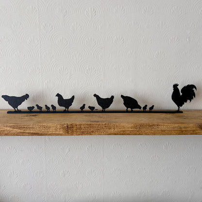 A black recycled plastic topper piece featuring a rooster and several chickens and chicks, displayed on a rustic wooden shelf against a white textured wallpaper background.