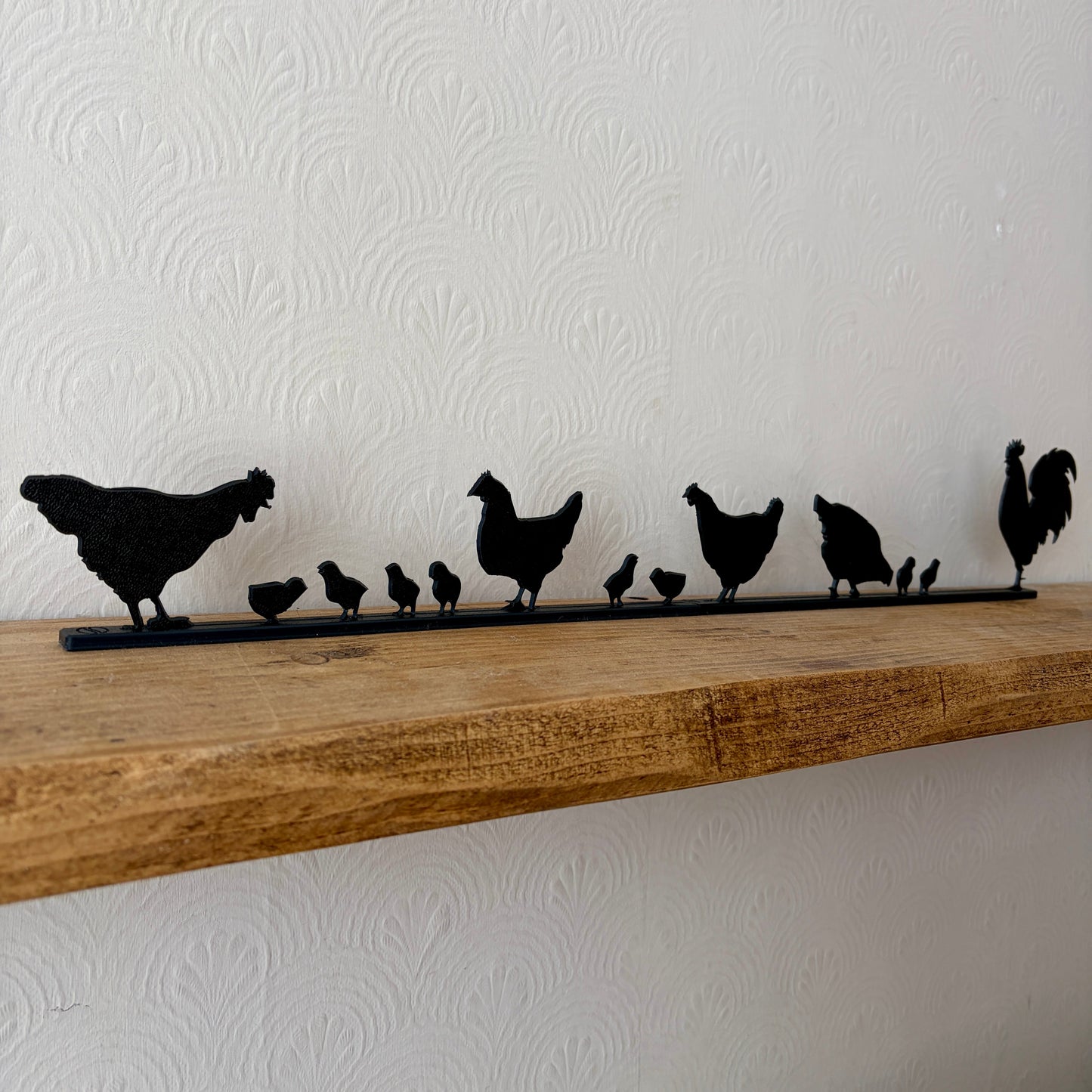 A black recycled plastic topper piece featuring a family of chickens and chicks, placed on a rustic wooden shelf against a white textured wall.