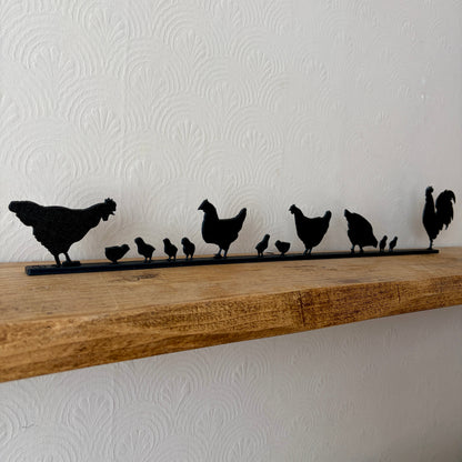 A black recycled plastic topper piece featuring a family of chickens and chicks, placed on a rustic wooden shelf against a white textured wall.