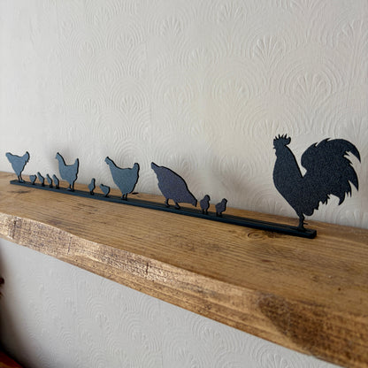 A black recycled plastic topper piece featuring a rooster and several chickens and chicks, mounted on a rustic wooden shelf against a textured off-white wall.