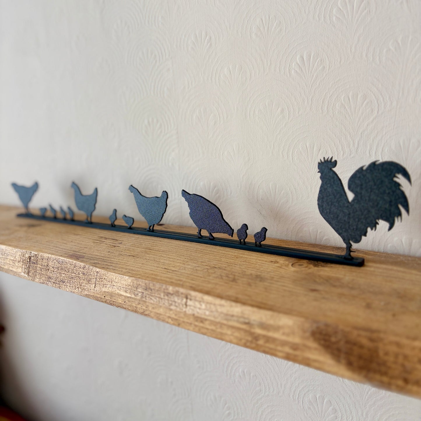 A dark recycled plastic topper piece featuring a rooster and several chickens and chicks, displayed on a rustic wooden shelf against a textured off-white wall.