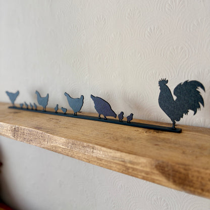 A dark recycled plastic topper piece featuring a rooster and several chickens and chicks, displayed on a rustic wooden shelf against a textured off-white wall.