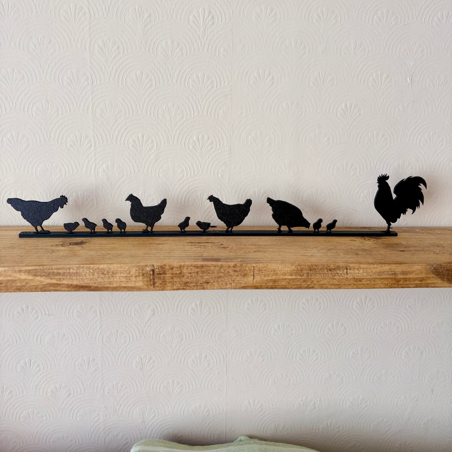 A black recycled plastic silhouette topper featuring a family of chickens and a rooster, displayed on a rustic wooden shelf against a textured off-white wall.