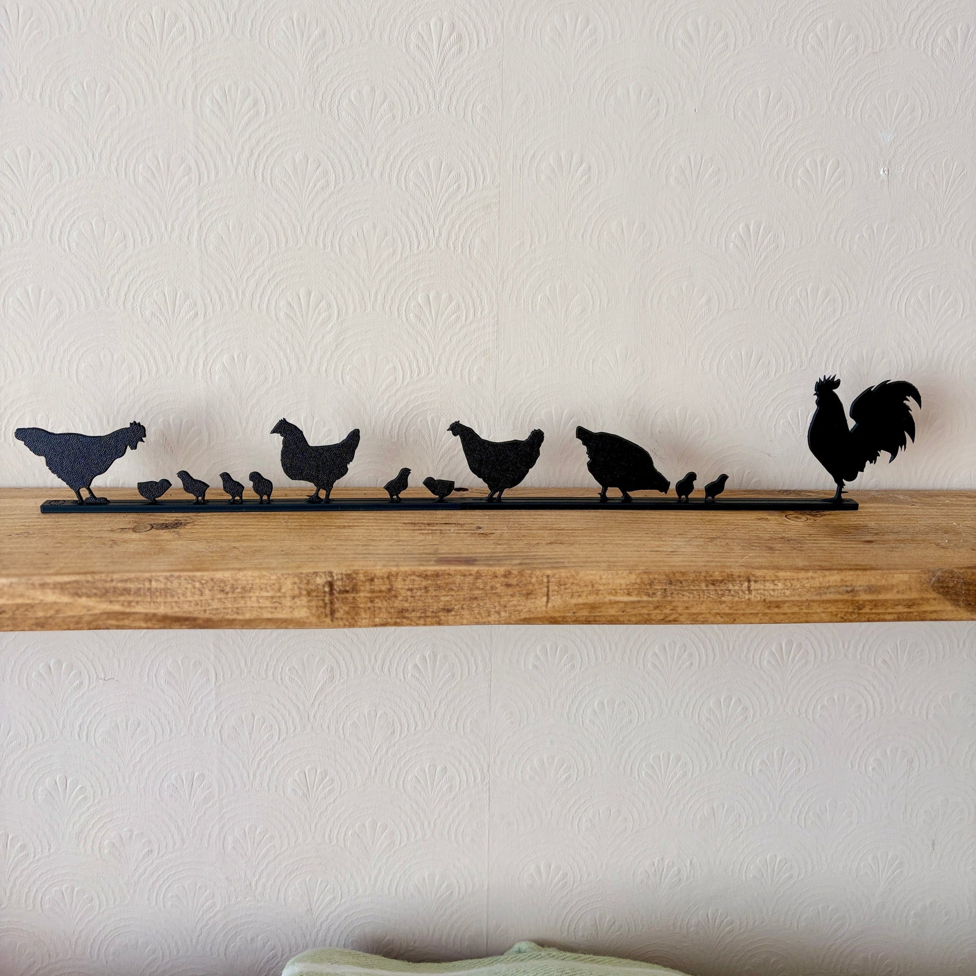 A black recycled plastic silhouette topper featuring a family of chickens and a rooster, displayed on a rustic wooden shelf against a textured off-white wall.