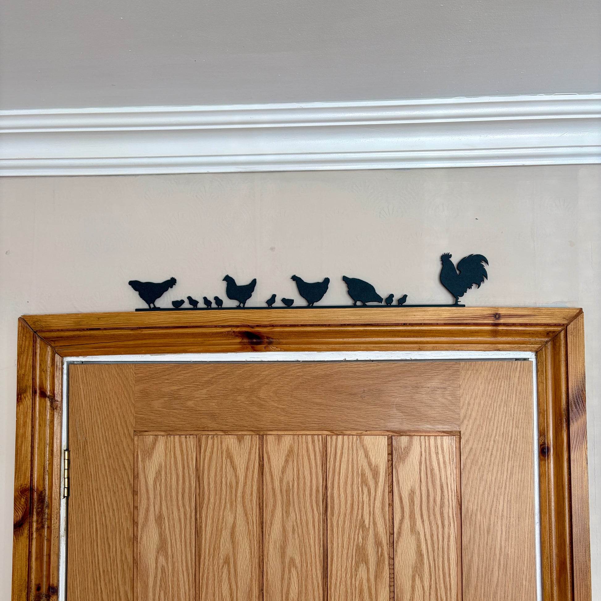 A black recycled plastic silhouette wall decor piece featuring a rooster and four chickens and their chicks, mounted above a wooden door frame.