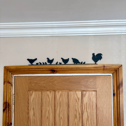 A black recycled plastic silhouette wall decor piece featuring a rooster and four chickens and their chicks, mounted above a wooden door frame.
